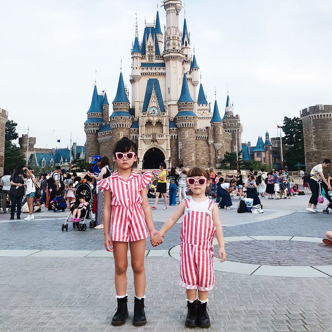 Two girls in matching striped outfits and sunglasses holding hands in front of the castle at Tokyo Disneyland