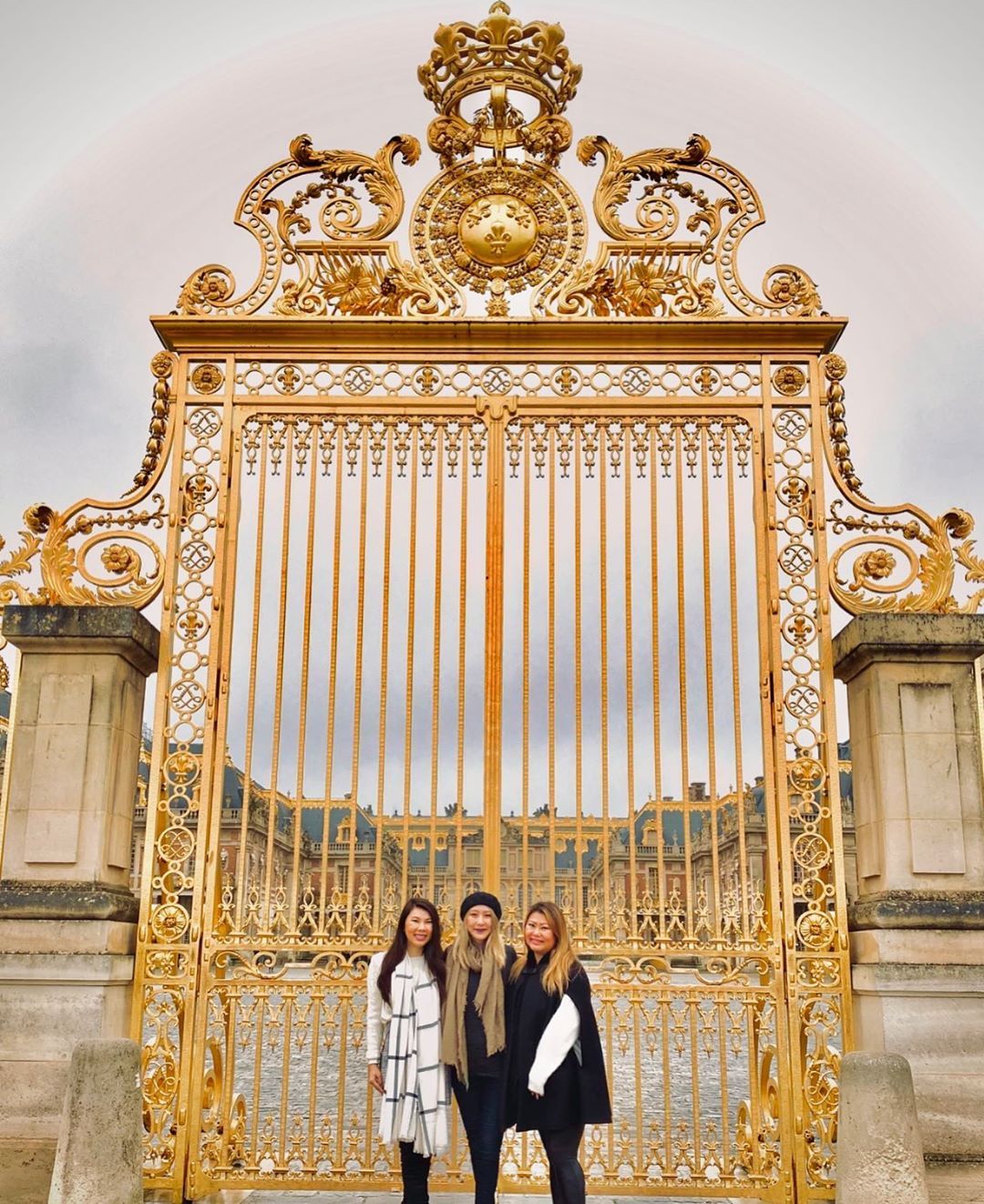 Three women posing in scarves and coats in front of the gate of the Palace of Versailles
