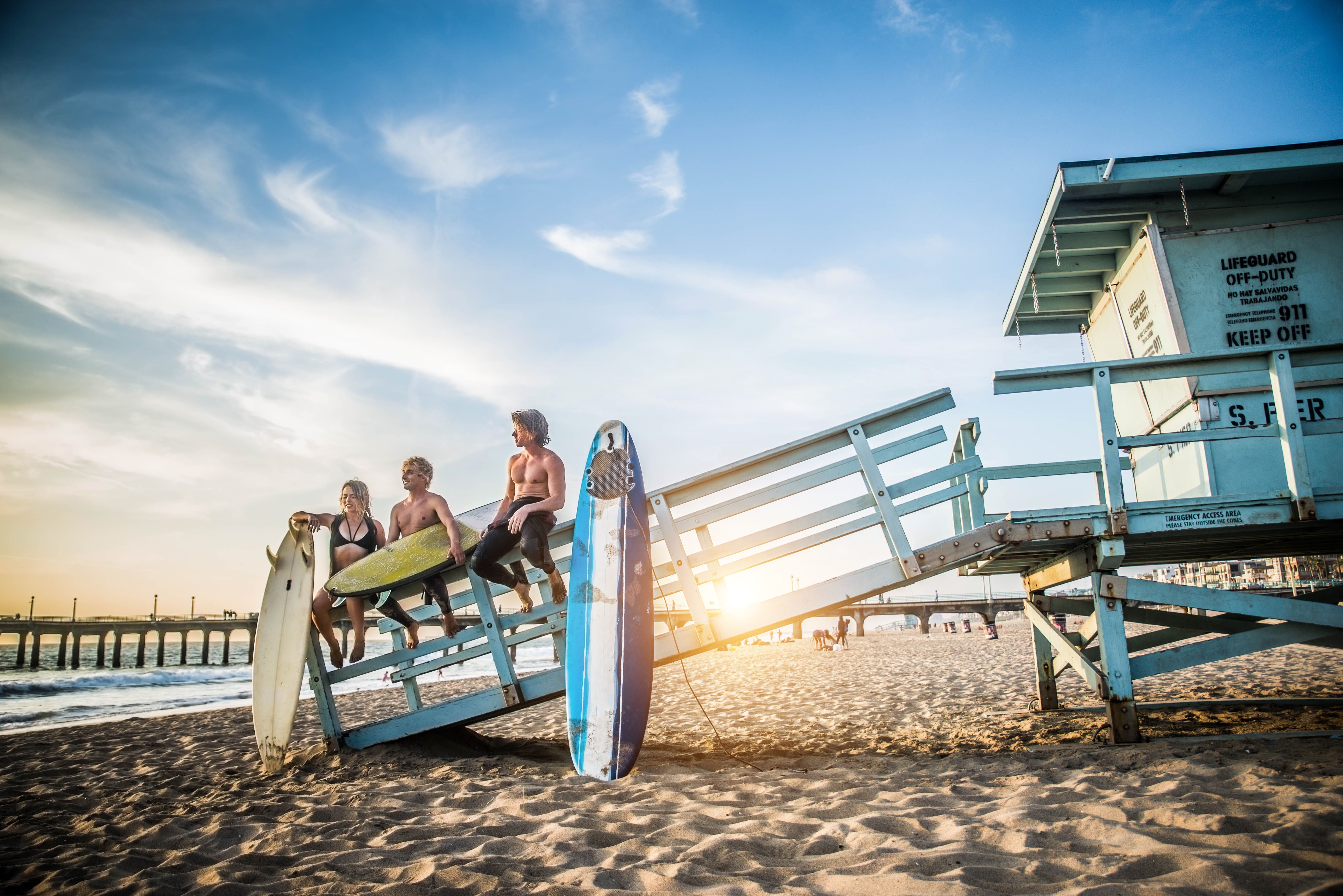 Surfers on a Los Angeles beach sitting on a walkway while holding their surfboards as the sun sets behind them