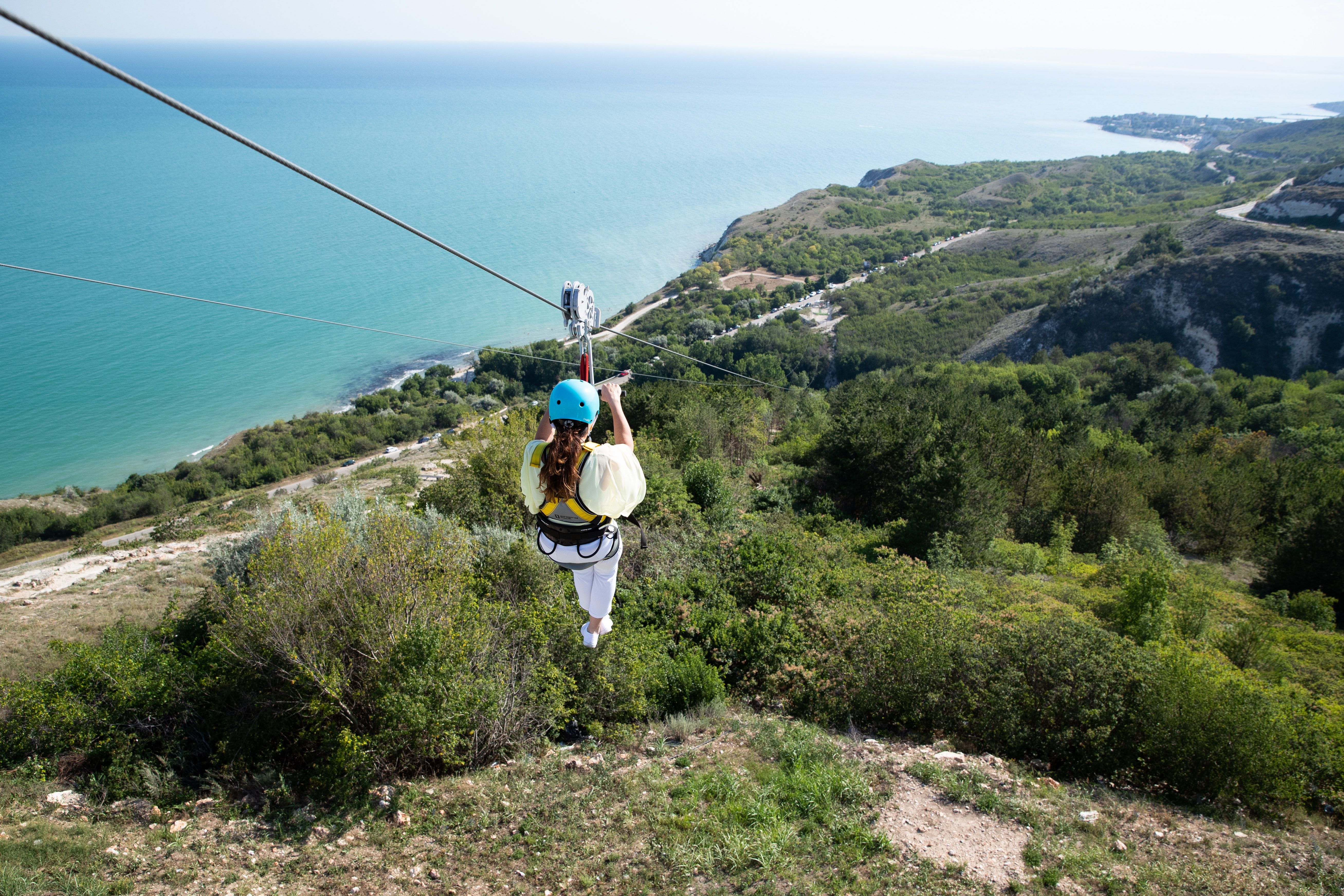 A lady ziplines over a valley by the sea.