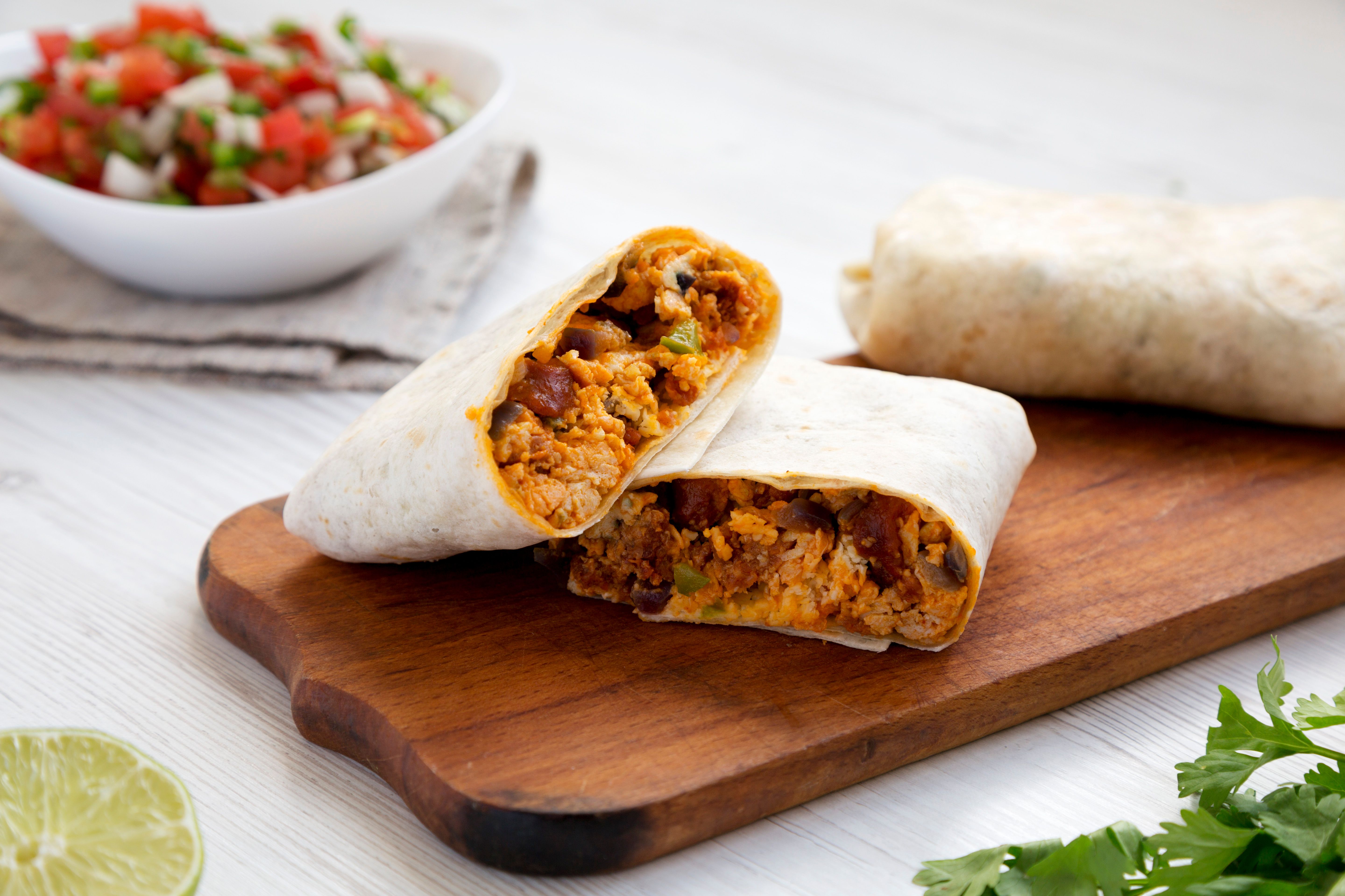 Homemade chorizo breakfast burritos on a rustic wooden board on a white wooden surface, low angle view.