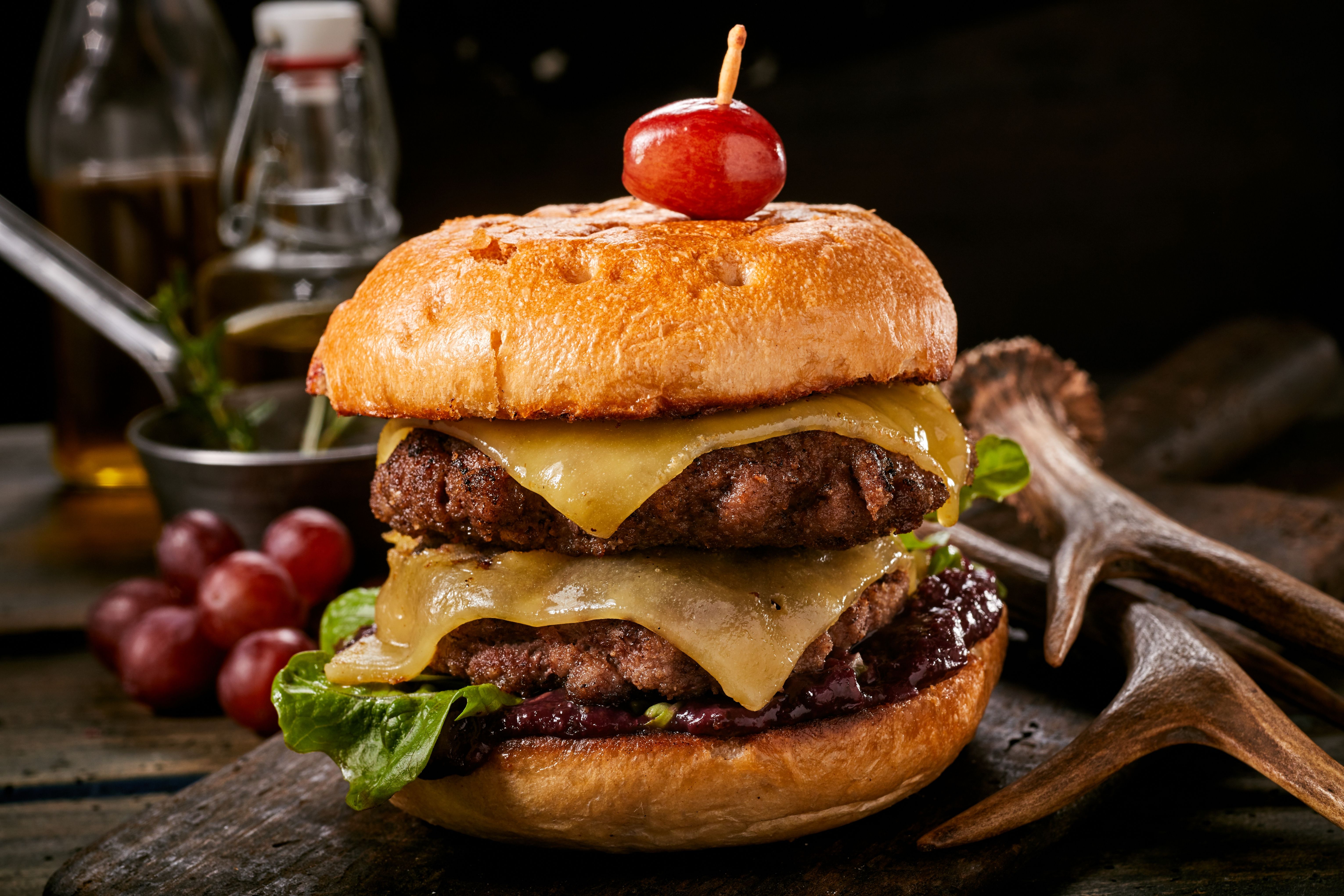 Delicious double venison cheeseburger with olives and melting cheese on a toasted crusty bun served on a rustic wooden table alongside deer antlers