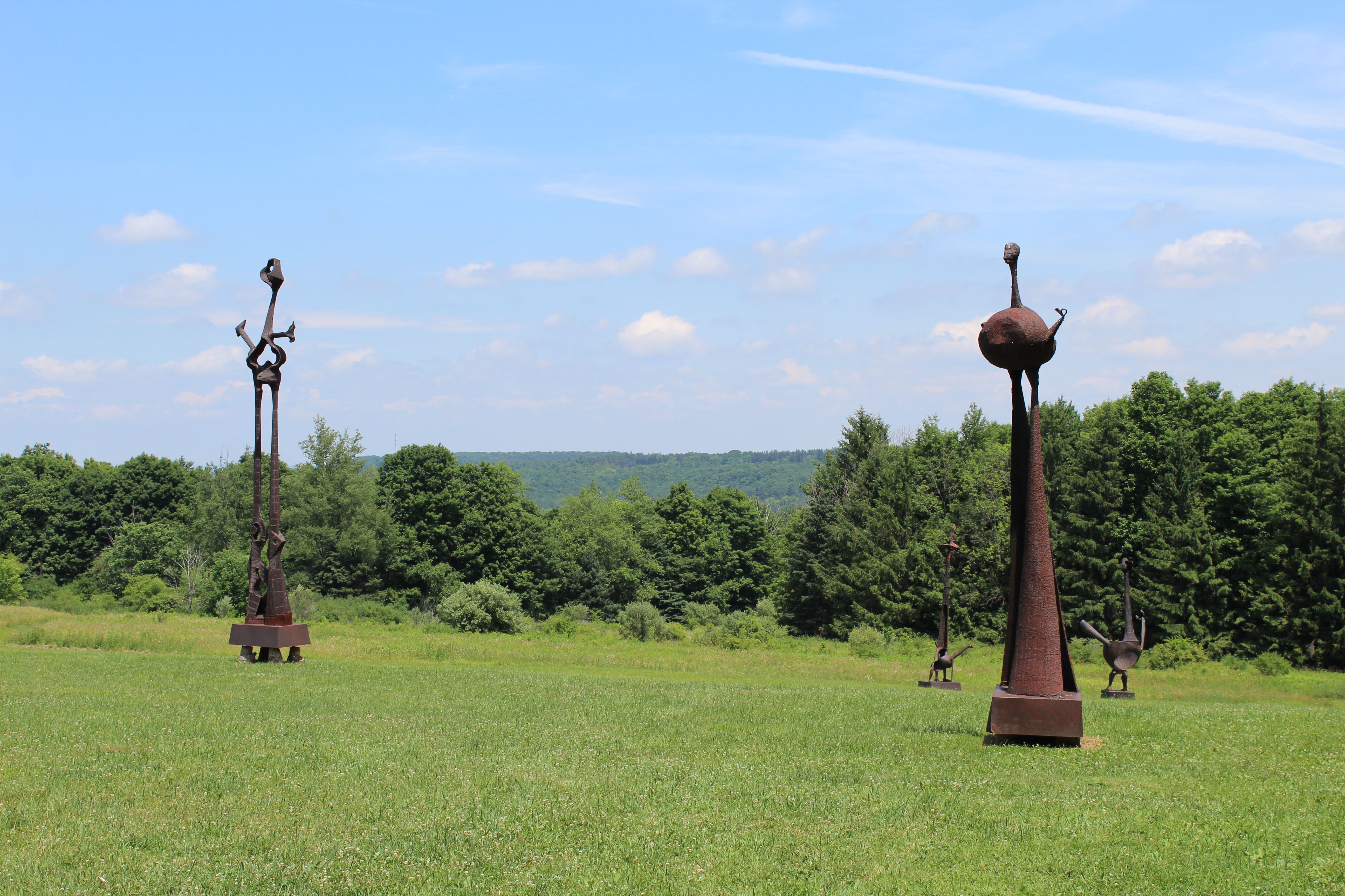 A photograph of Griffis Sculptue Park mid-day in relatively sunny weather. Multiple steel sculptures are displayed in a field, with woods in the background.