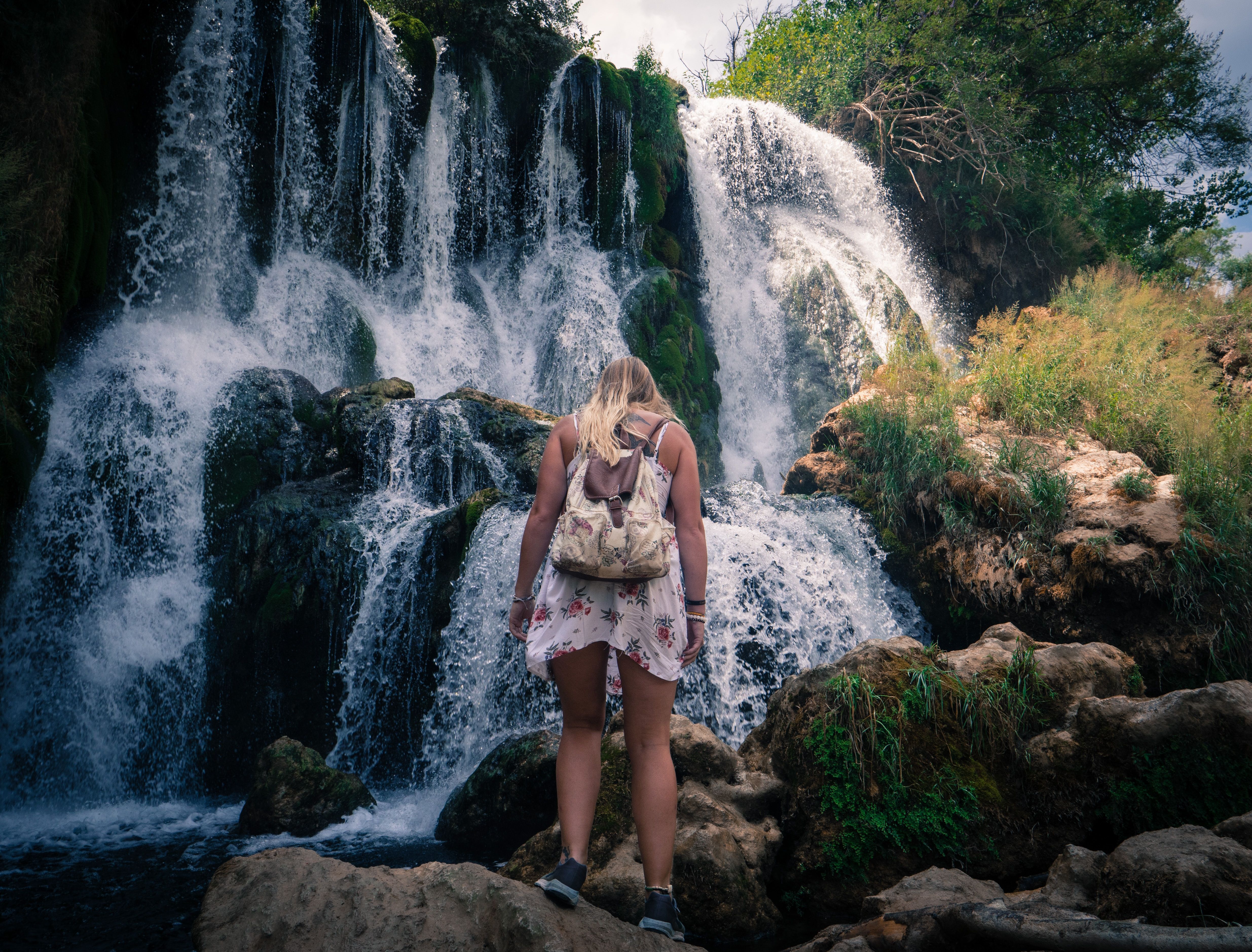 Mulher vestindo uma mochila fica na frente de uma cachoeira alta 
