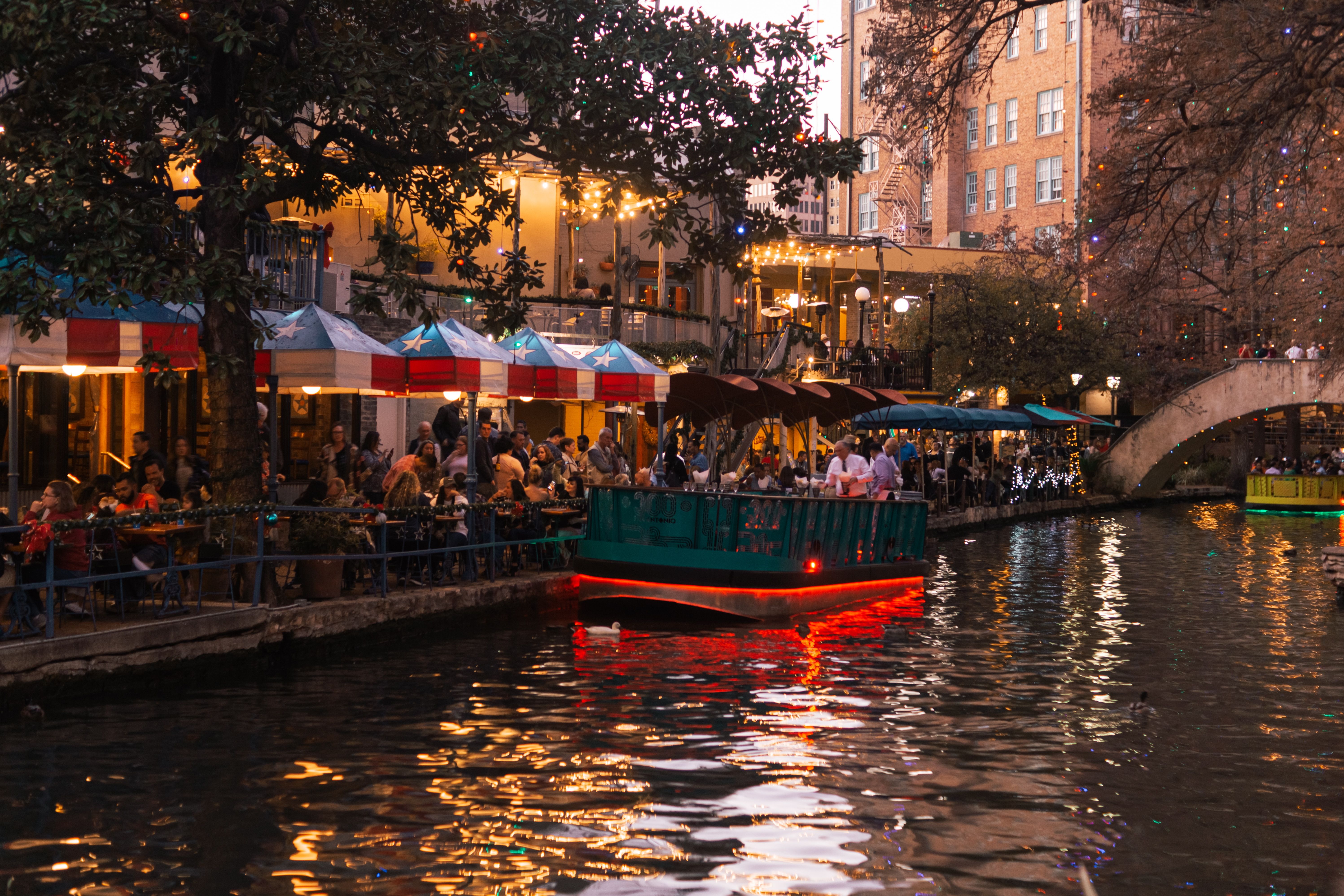 Boat In The Water Of A Canal With People Dining Along The Shore In San Antonio, Texas