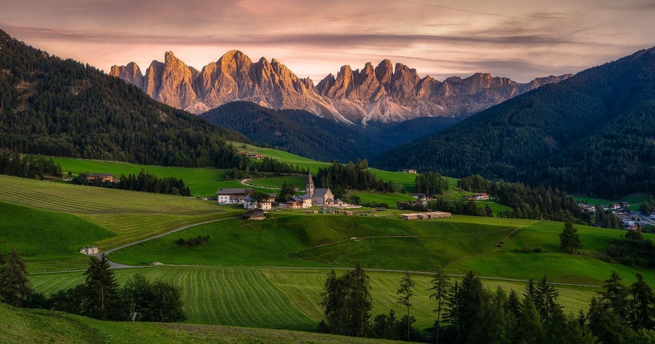 A scenic view of green grassy hills, forests, and the Dolomite Mountains in Italy