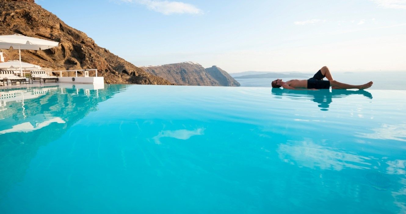 Man Relaxing On The Edge Of An Infinity Pool Overlooking Santorini Sea 