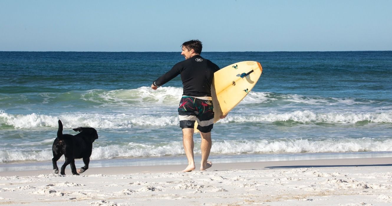 A man with a surfboard and a black dog on a white-sand beach in Florida, USA