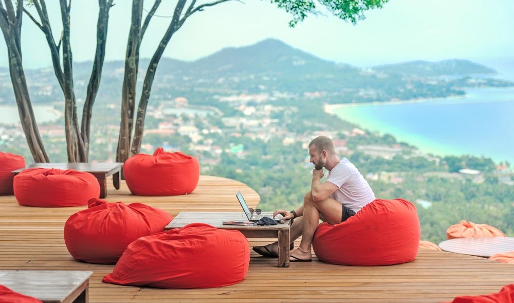 A man sitting on a red cushion working remotely with his laptop