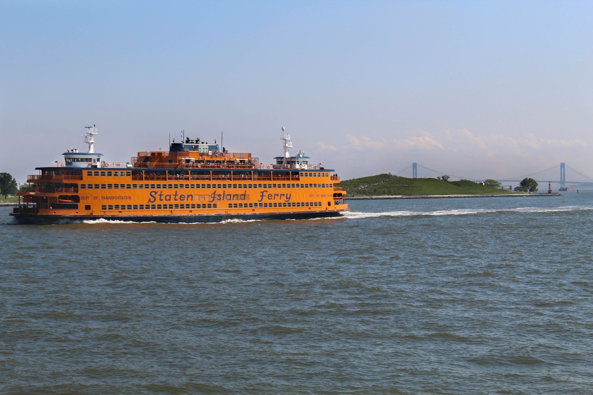 Staten Island Ferry in New York City traversing the water with green terrain and a bridge in the background