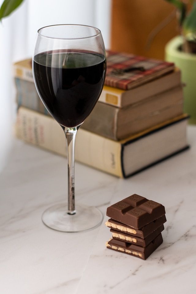 A glass of red wine next to some chocolate with several books behind them, all on a marble table