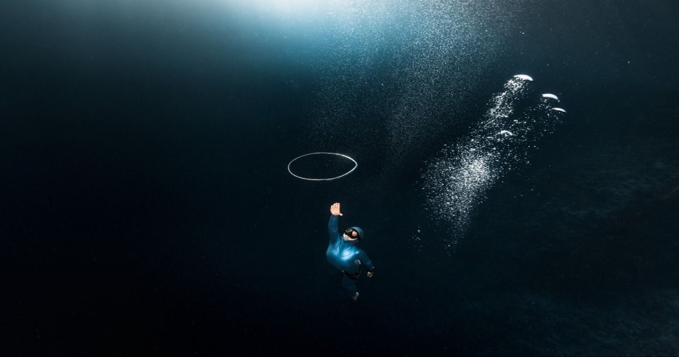 A woman freediver in a blue freediving wetsuit chasing a bubble ring undewater