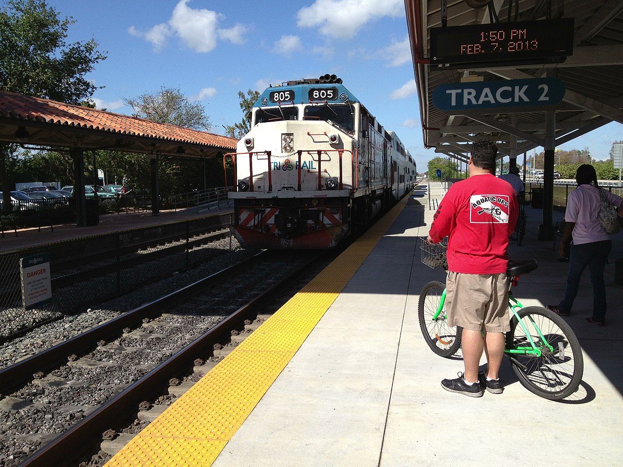 An Amtrak train nearing a stop on a sunny day with a man holding his bike at the platform