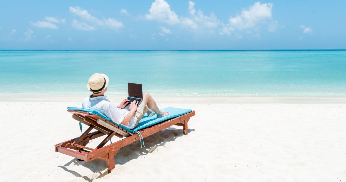 A man working as a digital nomad on a laptop computer on a white-sand beach in front of the turquoise ocean