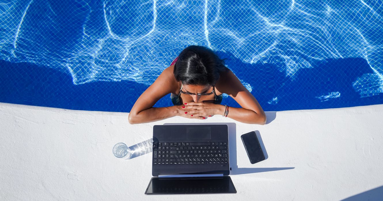 A woman working as a digital nomad on a laptop computer on the side of a pool