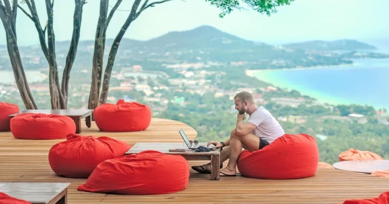 A digital nomad working on a laptop while sitting on a red bean bag on a wooden deck that overlooks a scenic beach, forest, and town
