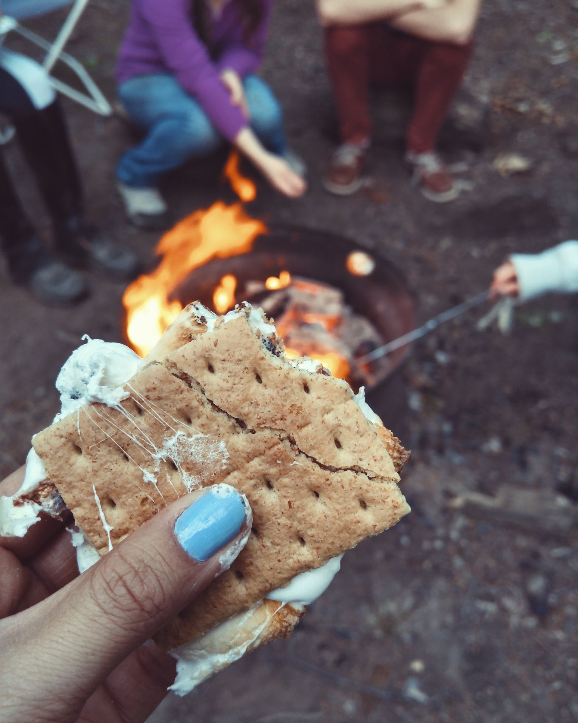 Woman holding a S'mores while others in the background roast marshmallows around a campfire. 