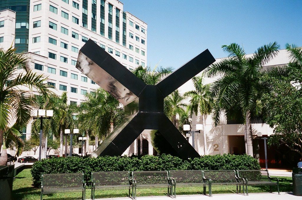 A sculpture of a black cross in front of Miami Dade College surrounded by benches.