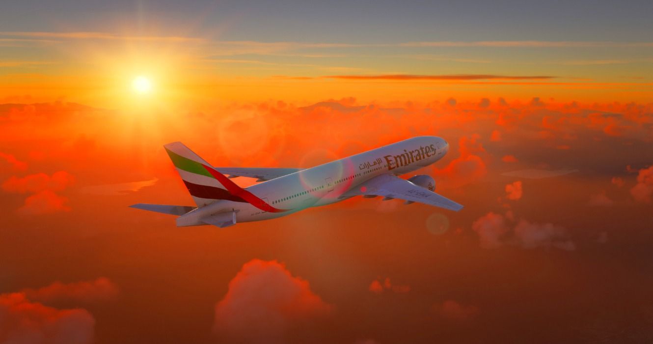 An Emirates Airlines Boeing 777 airplane flying through an orange sky at sunset or sunrise