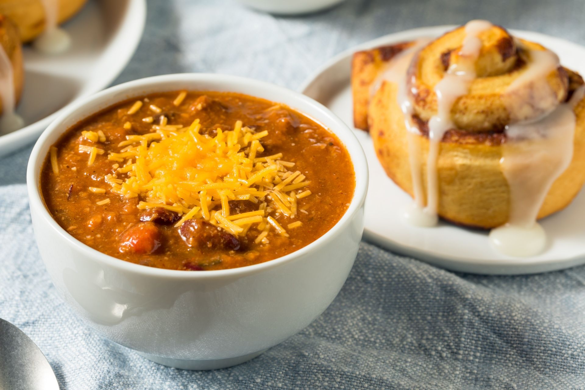 A steaming bowl of hearty chili next to a warm cinnamon roll on a plate.