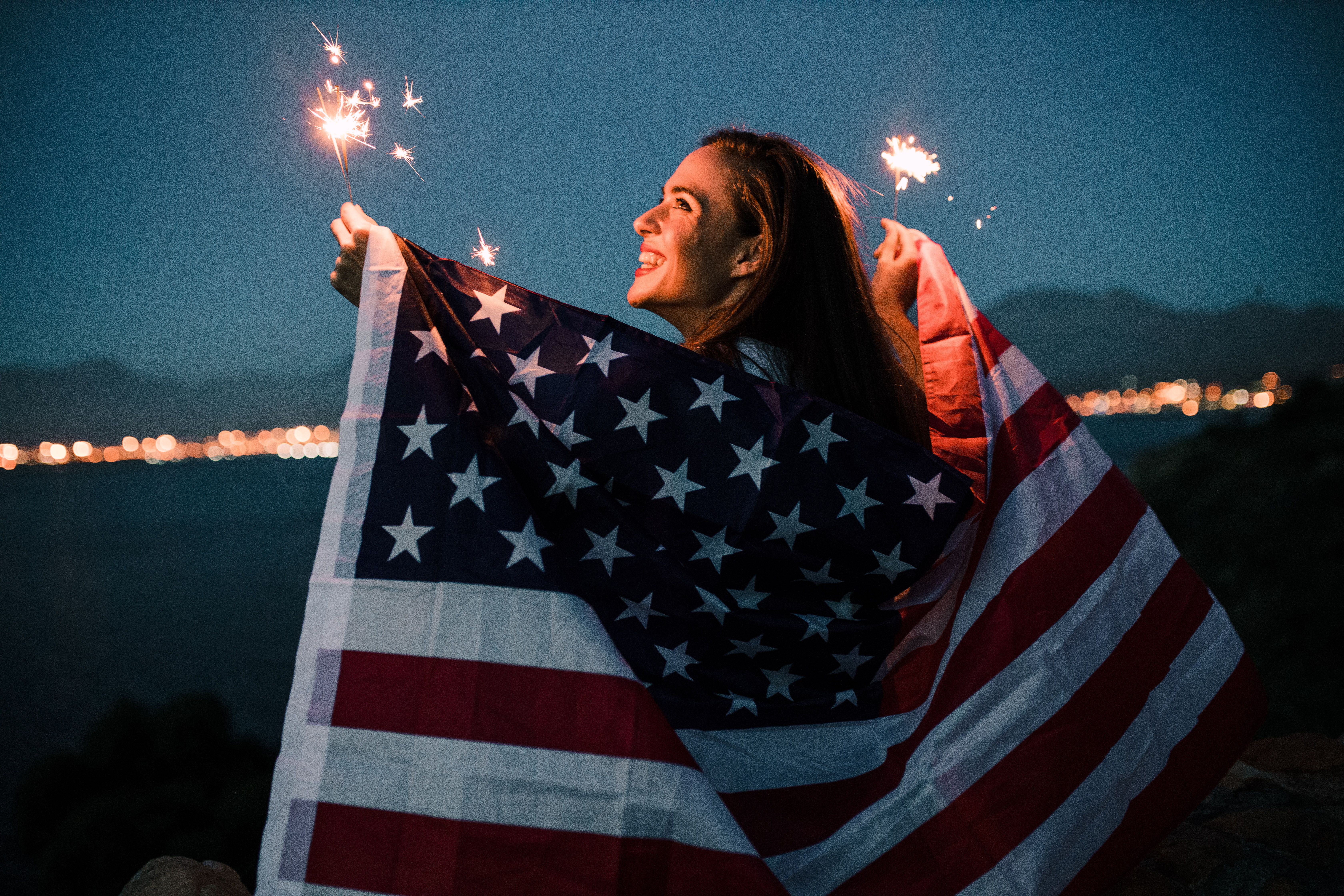 A woman draped in an American flag celebrates July 4th with a sparkler on a beach