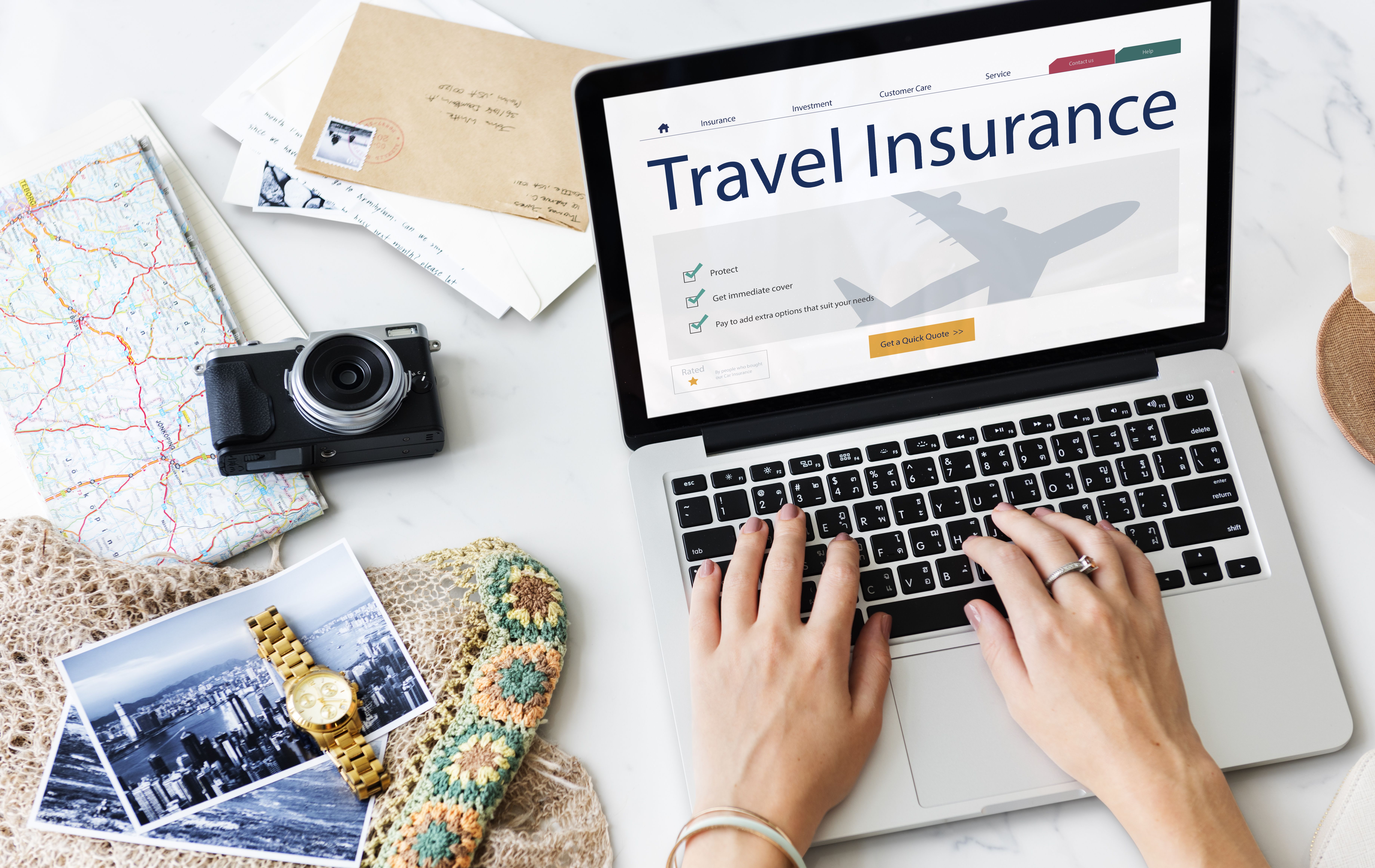 A person books travel insurance on a laptop with various travel essentials such as a camera next to them.