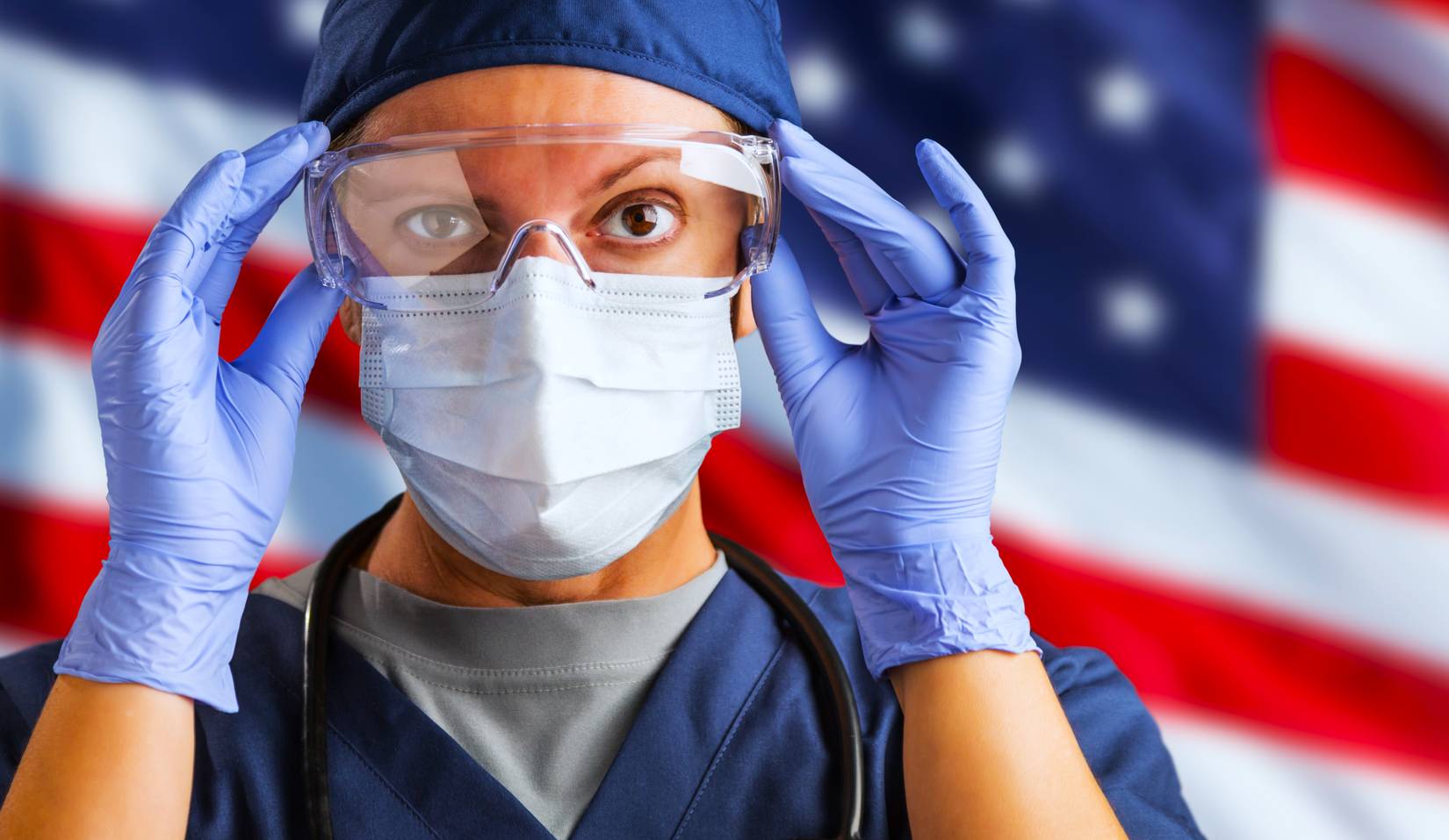 A nurse stands in front of a US flag while wearing a mask and PPE